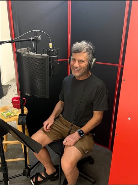 Steve Kaplan in headphones, smiling at the microphone in a recording booth, narrating the audiobook